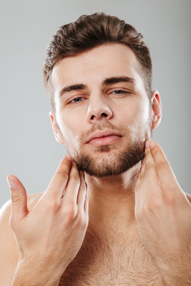 Close up portrait of a young bearded man looking at camera with hands on chin isolated over gray background