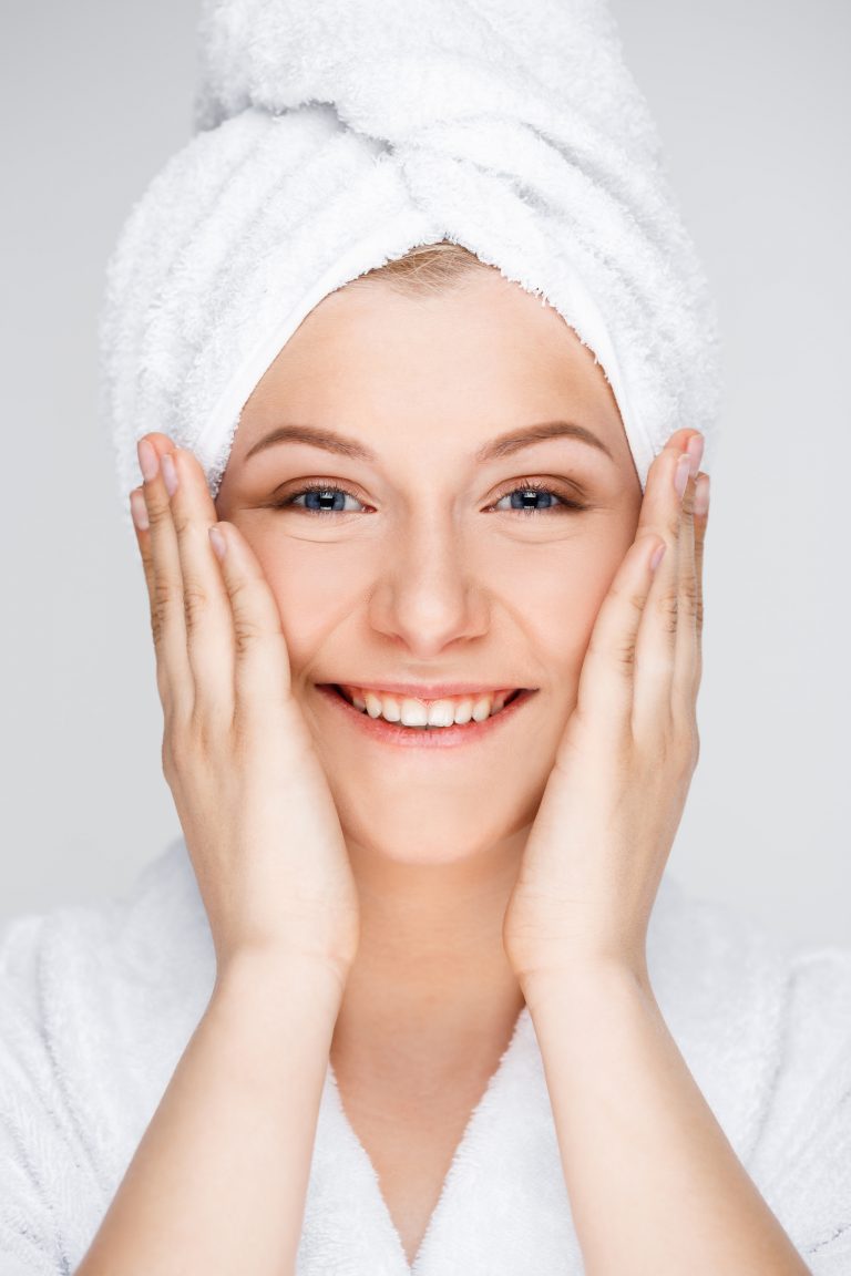Portrait of blonde young pretty girl in bathrobe with towel on head, smiling, looking at camera, hands on cheeks, over white background.